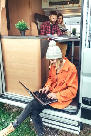 Female in jacket and woolen hat sitting on step of van with laptop, vertical
