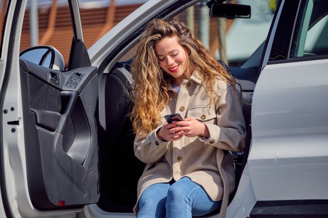 Smiling woman in coat browsing smartphone and sitting in car