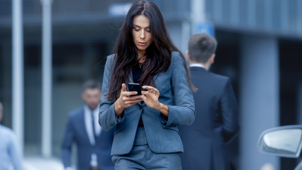 Business Woman in the Tailored Suit Walking on the Busy Big City Street in the Business District, Checks Her Smartphone.