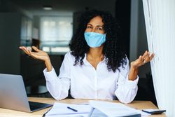 Woman in facemask with her arms up with documents and laptop in front of her
