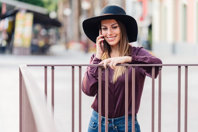 Charming woman in trendy outfit talking on smartphone and smiling at city street