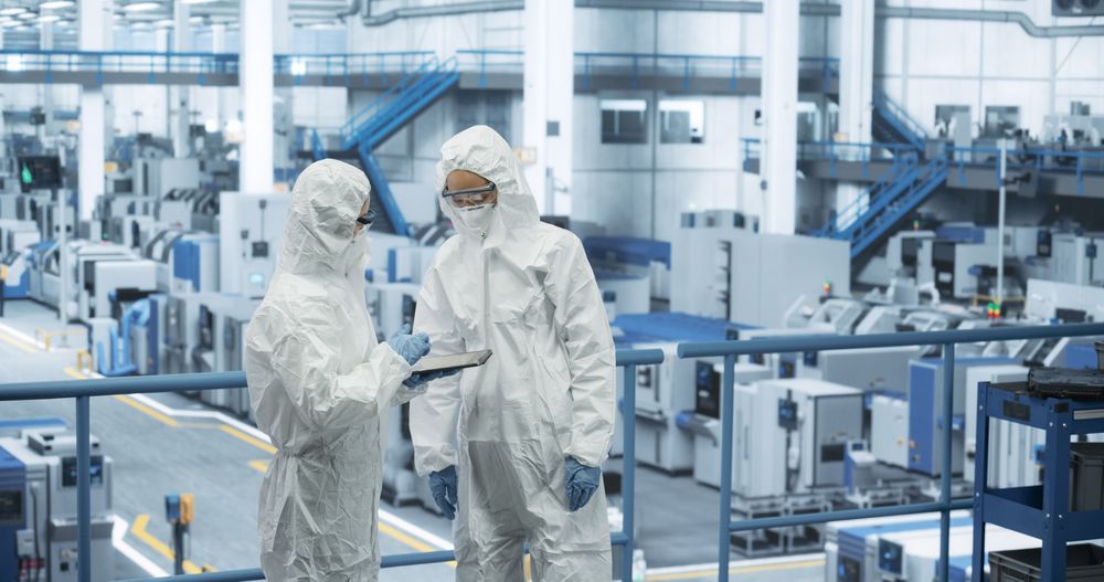 Two Female Technicians in Protective Coveralls Having a Conversation and Using Tablet Computer. Specialists Monitoring, Analyzing Conditions at a Modern Electronics Factory with Automated Robots