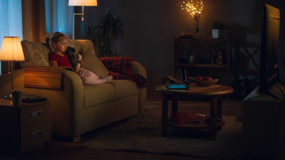 In the Evening Cute Little Girl is Sitting in Living Room on a Sofa Watching Television.