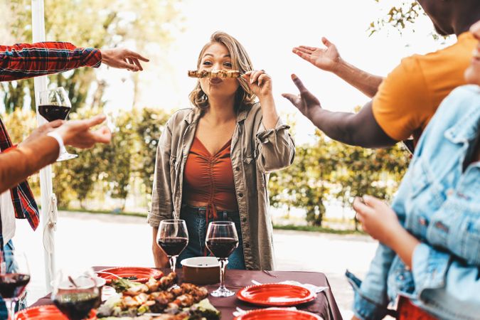 Friends having fun party with a woman making a funny face holding skewers grilled on barbecue