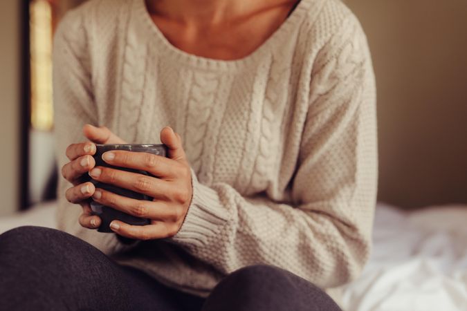 Close up of woman wearing sweater holding a cup of coffee while sitting on bed