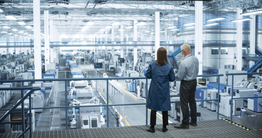 Mature Engineer and Female Scientist Using Laptop Computer and Talking in a Factory Facility with Equipment Producing Modern Electronic Components for Different Industries