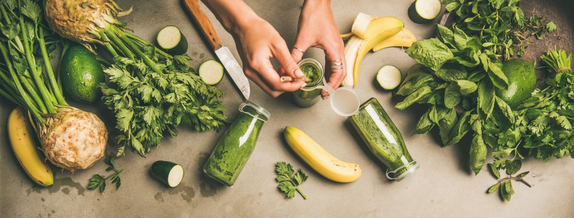 Smoothie ingredients with hands holding bottle, banana, zucchini, on grey table