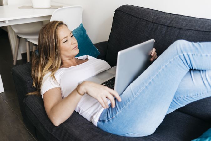 Long haired woman lying on sofa with laptop computer working remotely from home