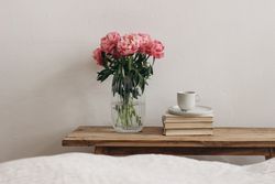 Pink peonies flower on glass vase on wooden table with pile of books and cup of coffee