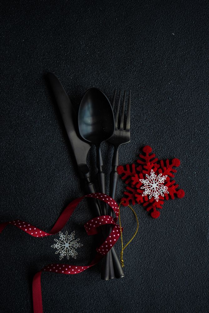 Dark knife and fork on dark background tied with festive red ribbon and snowflake ornament