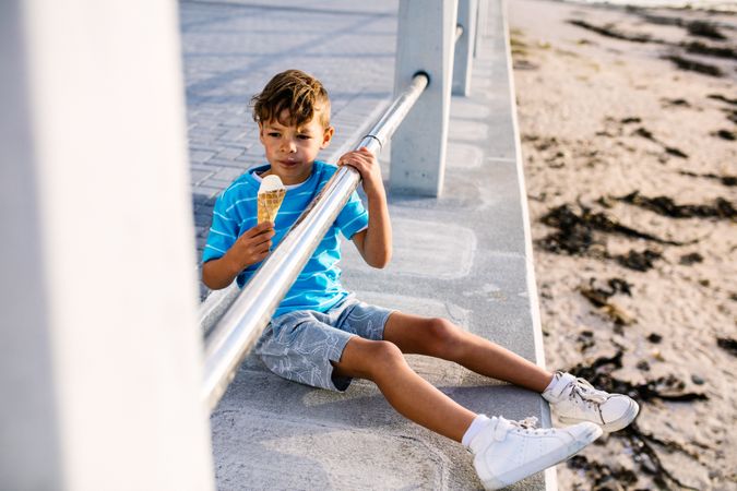 Boy eating an ice cream sitting near a seashore railing