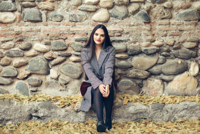 Female looking at camera in warm winter clothes sitting in front of rock wall in fall