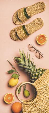 Sandals, glasses, pineapple and fruits in wicker bag, on pink background, vertical composition