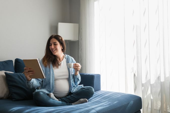 Cheerful pregnant woman drinking beverage on sofa at home