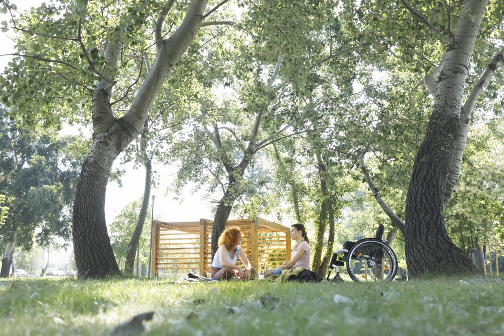 Two women sitting under trees in a park with wheelchair