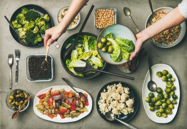 Flat-lay of vegetable salads, legumes, beans, olives, sprouts, hummus with woman’s hands
