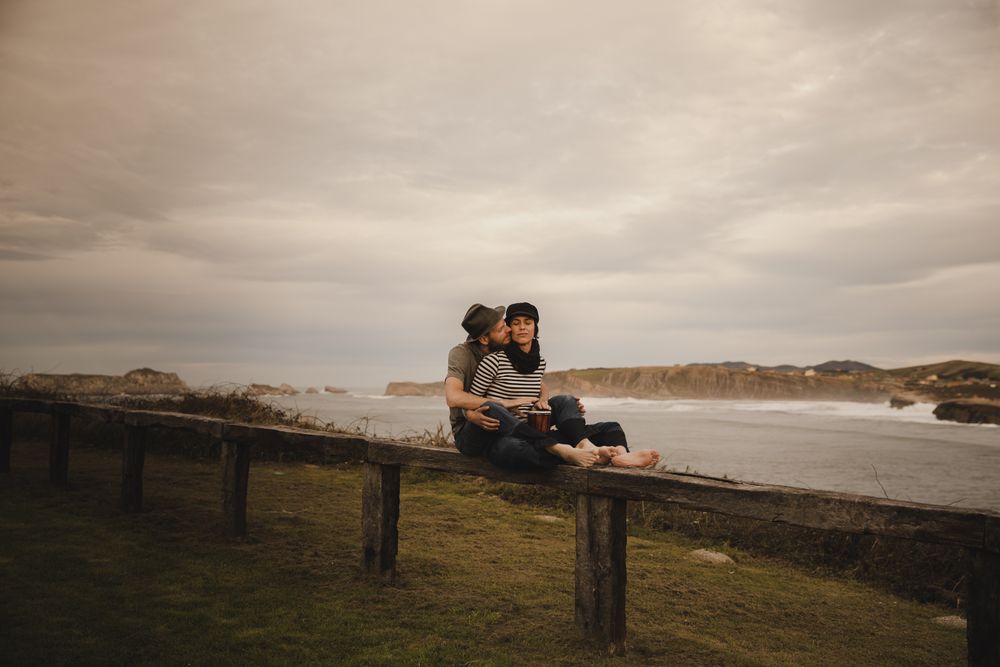 Young guy in hat near elegant woman playing on hand drum in cap on seat near coast of sea and cloudy sky