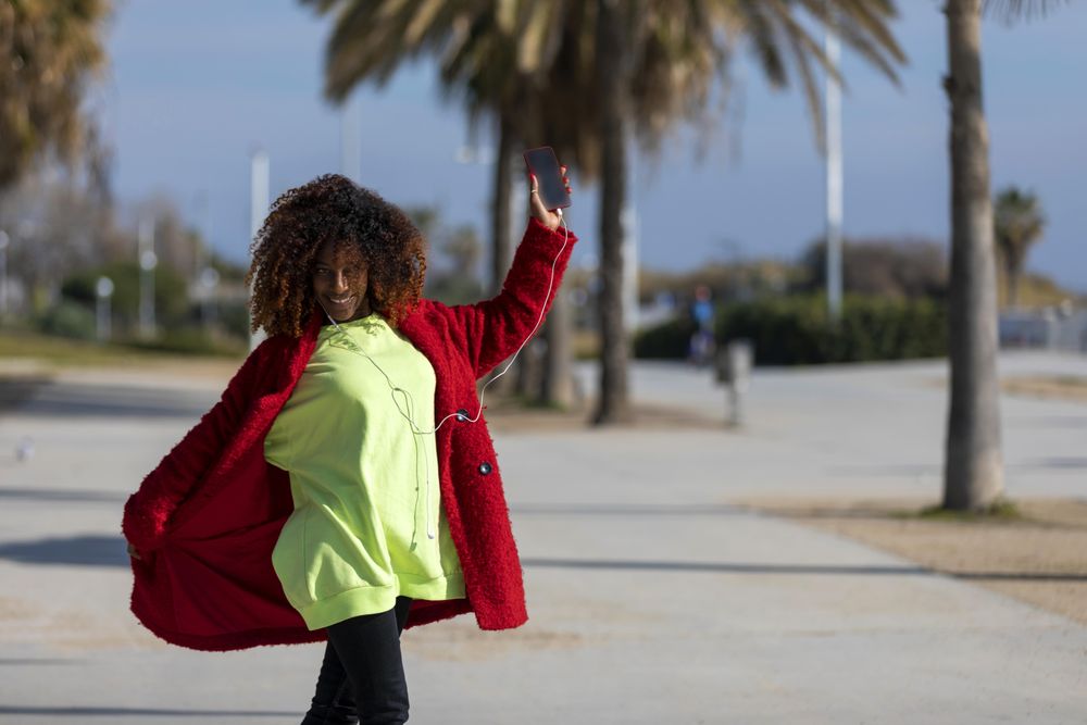 Smiling female in colorful clothes dancing to music on phone on street with palm trees