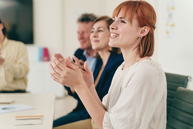 A group of colleagues applauds at the office desk, featuring a red-haired woman in the foreground and two other colleagues