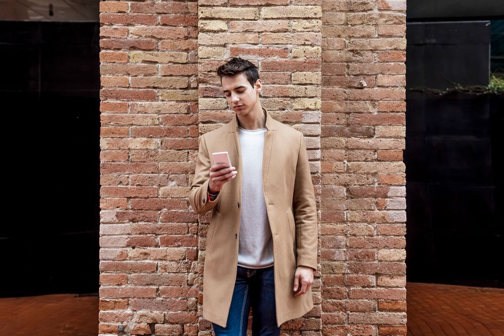 Front view of man in camel coat texting on phone while leaning on brick wall