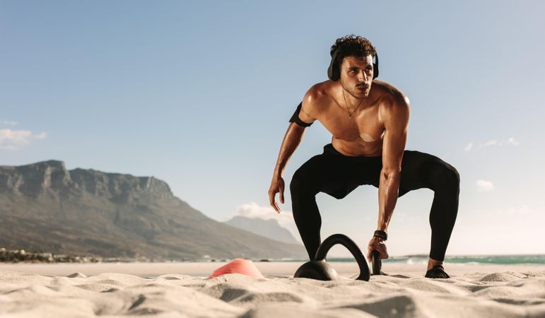 Man wearing headphones and doing kettlebell exercises on the beach
