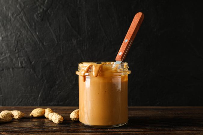 Glass jar with peanut butter and spoon, peanut on wooden table against dark background, space for text