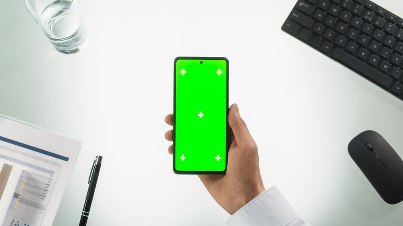 Top Down View of a Person Holding a Smartphone with Mock Up Green Screen Chromakey Display with Motion Tracker Placeholders. Office Desk with Wireless Computer Gadgets, Pen, Documents.