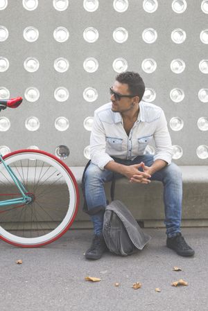 Relaxed male sitting with bike parked in front of patterned cement wall, vertical
