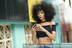 Young woman with afro hair checking phone near a modern colorful building 0V26Y0