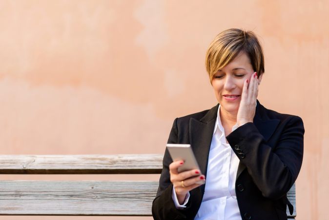 Bemused woman in blazer sitting on bench checking phone in front of peach wall