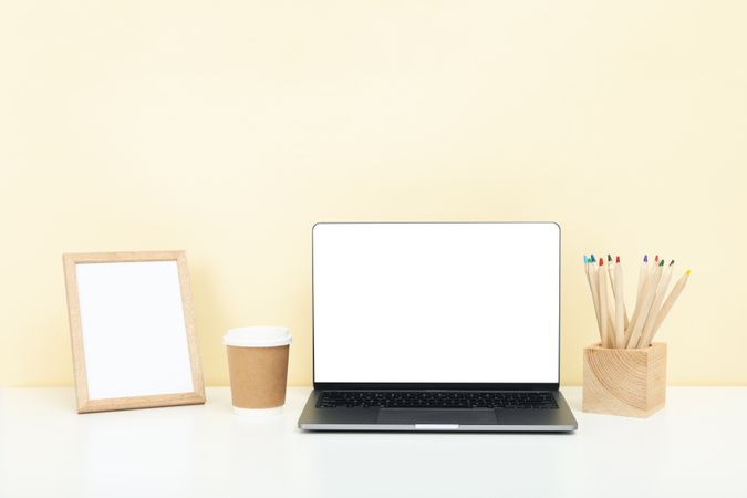 Open laptop with blank mockup screen in cream room with pencils, to go cup and to go drink