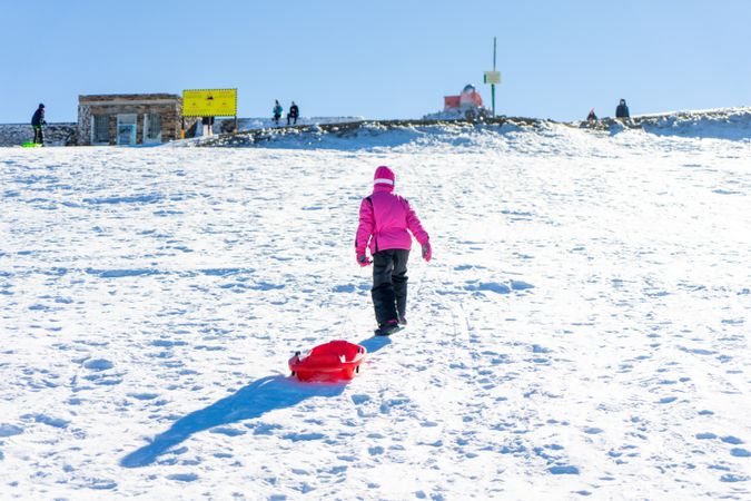 Child in pink snow suit pulling red sled behind her up a snowy hill