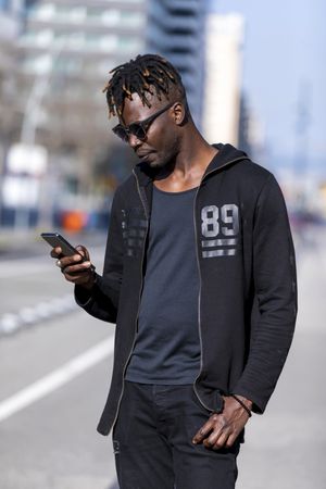 Side view of Black man standing on the street wearing casual clothes and sunglasses while using a mobile phone against blue sky