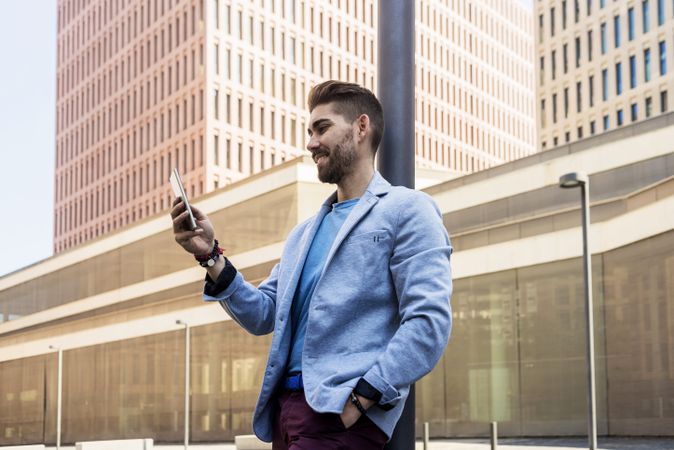 Man in blazer leaning on pole with one hand in pocket checking phone