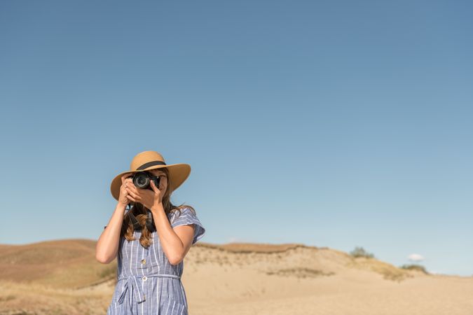 Back view of adult woman in straw hat and dress with camera taking photo on sand dune