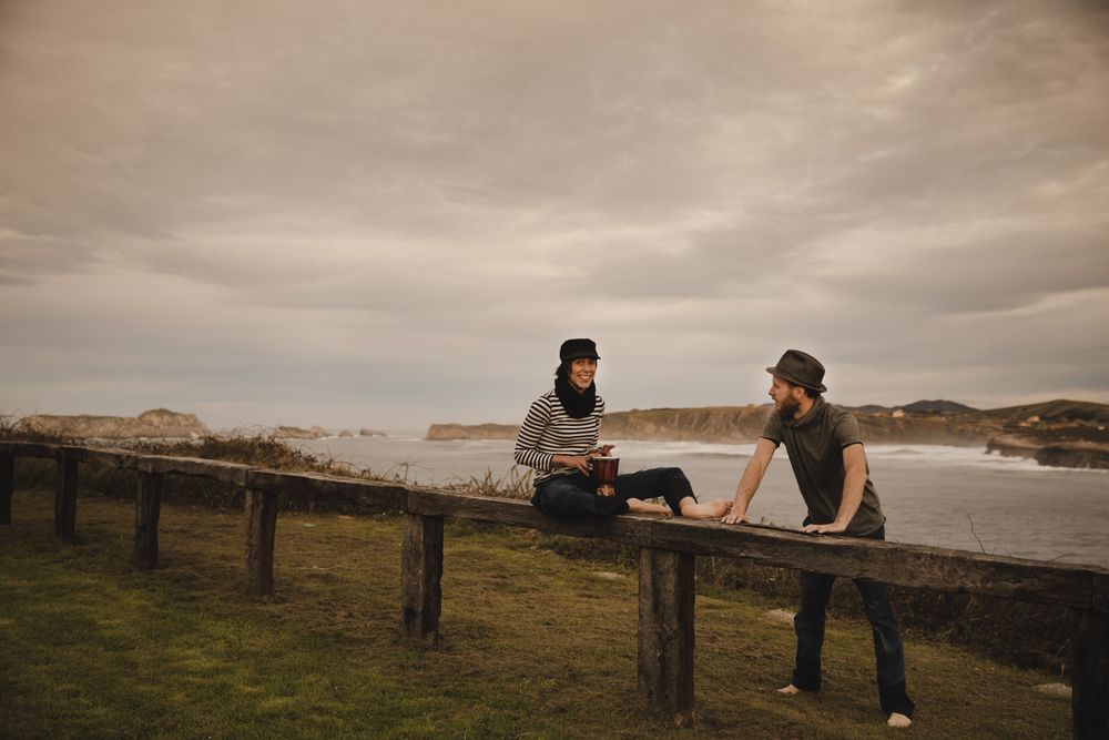 Young guy in hat near elegant woman playing on hand drum in cap on seat near coast of sea and cloudy sky
