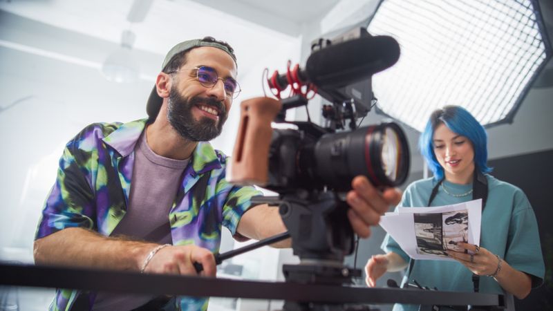 Young Male Cinematographer With Beard And Colorful Shirt Operates A Camera While A Female Colleague With Blue Hair Reviews Documents In A Well-lit Studio, Showcasing Teamwork In Movie Production.