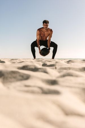 Man squatting with medicine ball on sand against blue sky