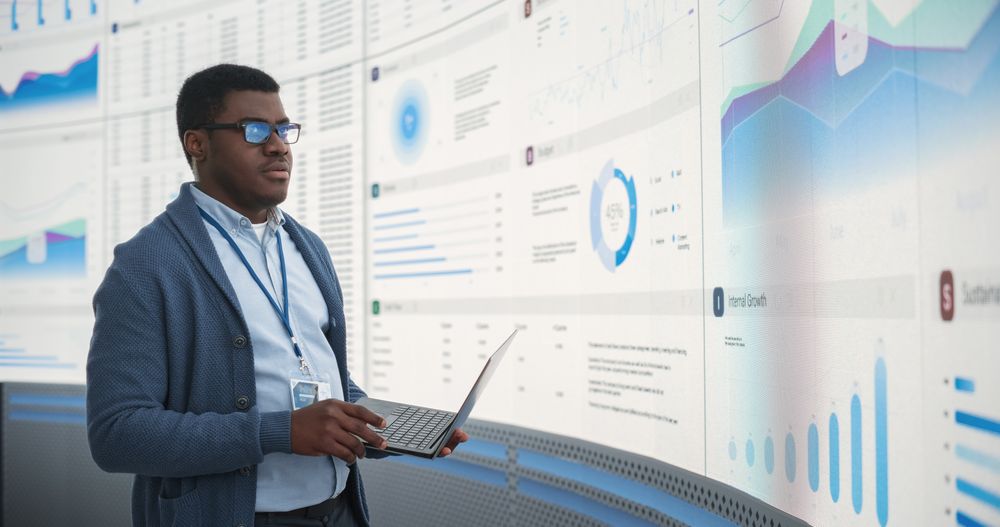 Confident Black Businessman Holding Laptop And Reviewing Financial Reports On Big Digital Screen In Monitoring Office.