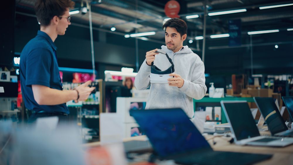 Young Man Having a Conversation with a Salesperson in a Electronics Store while Trying Out a Virtual Reality Device.