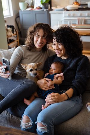 Two women with dog and baby on sofa making video call, vertical
