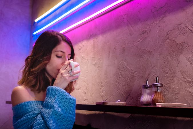 Positive woman with cup at table near wall with neon lamps
