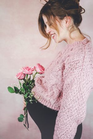 Side view of smiling pregnant woman in knit sweater holding pink rose bouquet