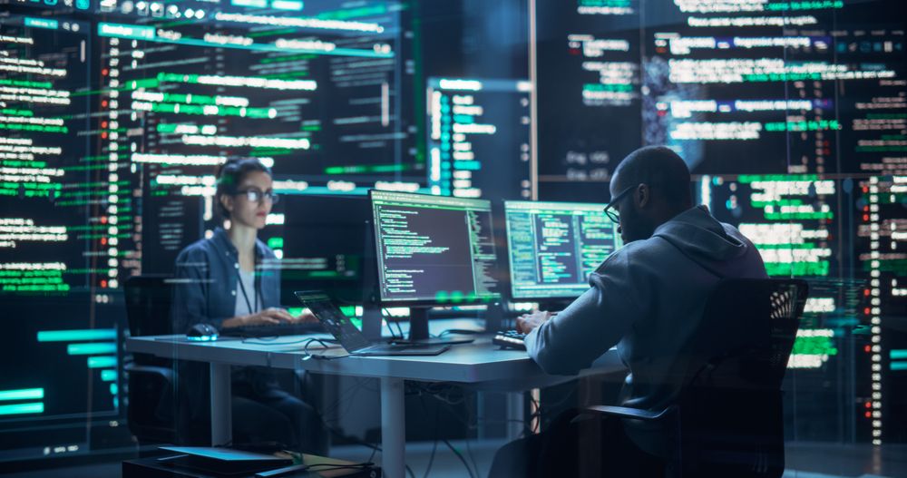Portrait of Two Programmers Working in a Monitoring Control Room, Surrounded by Big Screens Displaying Lines of Programming Language Code.
