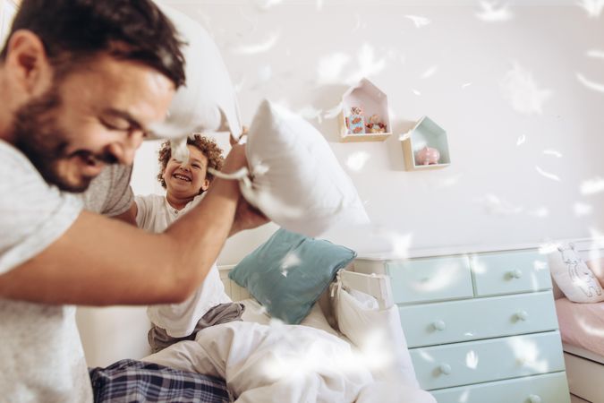 Cheerful man pillow fighting with son sitting on bed at home