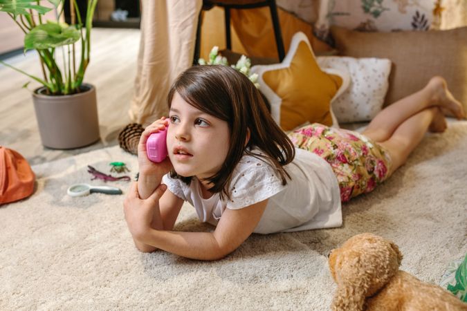 Little girl talking with toy phone lying over rug surrounded by cushions and plants in teepee