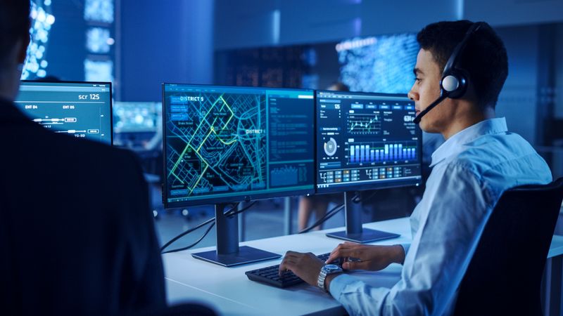 Male Professional in a High-tech Control Room Analyzes Real-time Data on Dual Monitors, Enhancing Cybersecurity and Operational Strategies. the Environment Fosters Innovation and Precision.