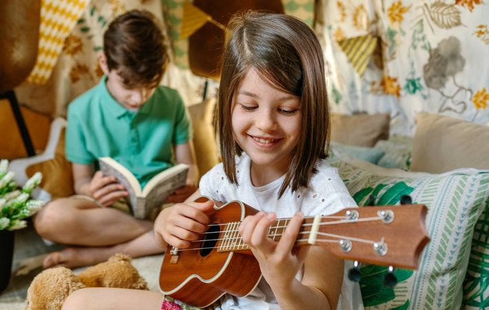 Happy smiling little girl playing ukulele while boy reading book on handmade shelter tent
