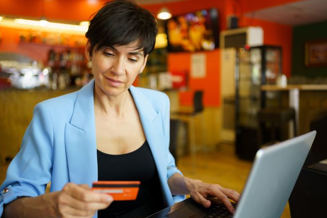 Female in trendy blue jacket sitting in cafe with laptop and credit card