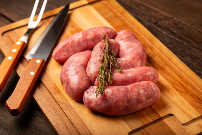 Raw sausages on wooden board with rosemary and utensils